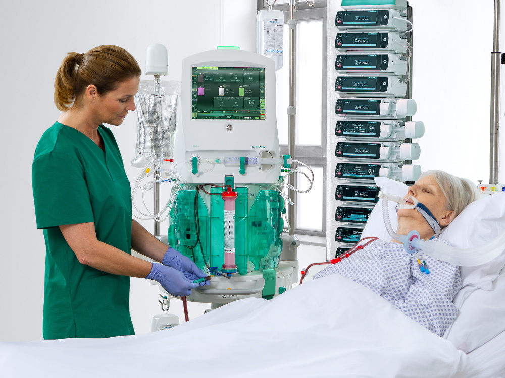 A middle-aged woman lies in the hospital bed. A nurse stays next to her and infuse some medication via medical device